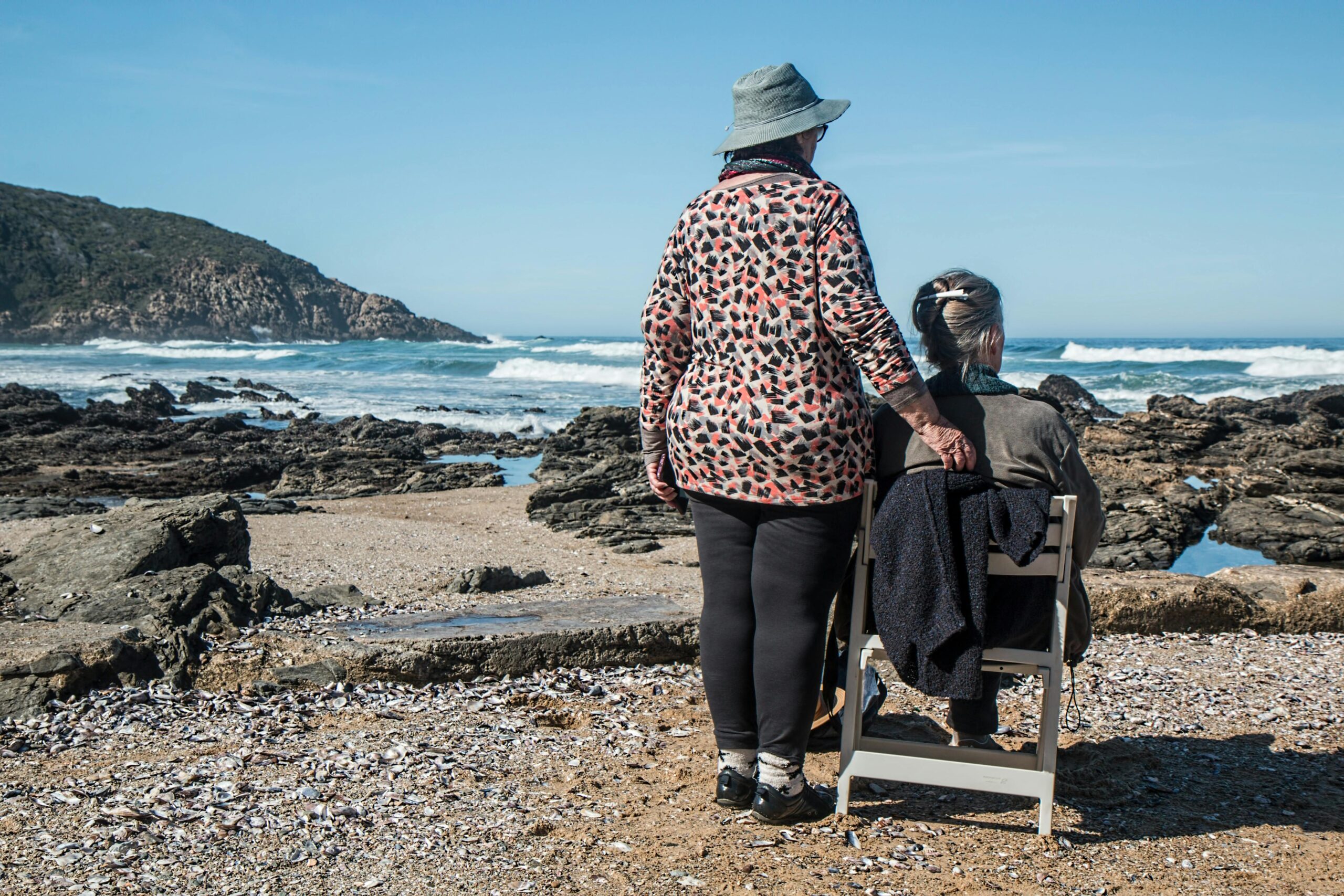 Duas idosas observando o mar numa praia rochosa, simbolizando o alívio financeiro das pensões de agosto com nova retenção de IRS.